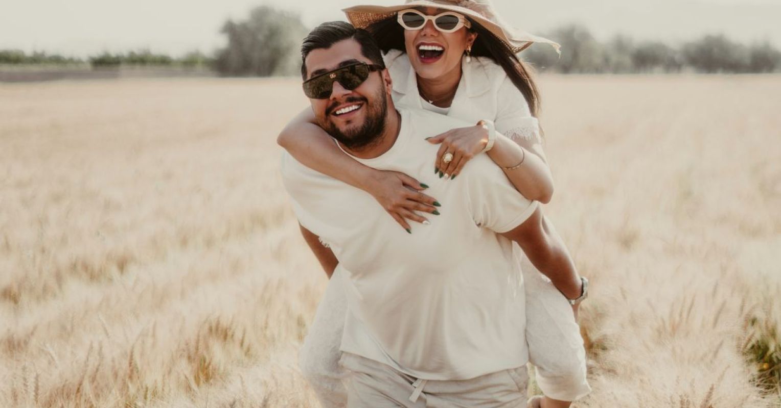 a man in sunglasses carrying a woman with a hat on his back through a wheat field 4x3 1